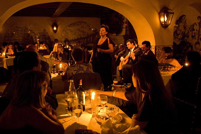 Gruppo di persone assiste, seduto ai tavoli di una casa di fado, a uno spettacolo.